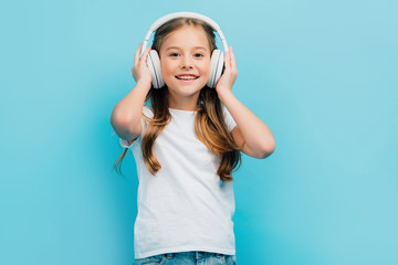 girl in white t-shirt touching wireless headphones while looking at camera isolated on blue