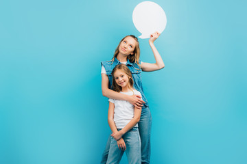 girl in white t-shirt and jeans looking at camera while mother holding thought bubble isolated on blue