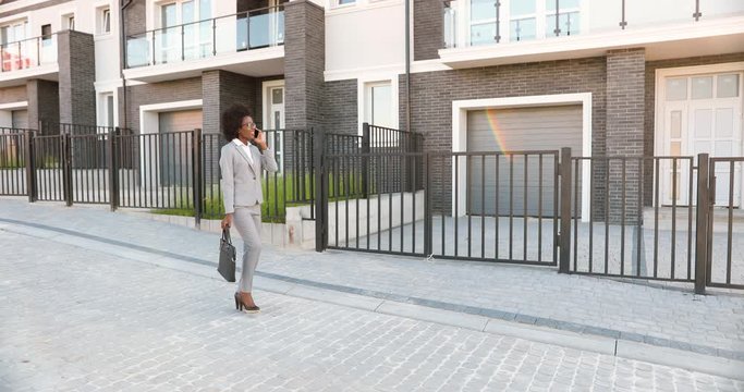 African American young stylish beautiful female real-estate agent walking the street at suborb neighborhood and talking on mobile phone. Businesswoman speaking on cellphone outdoor at outskirt.