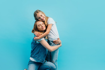 mother with closed eyes wearing denim clothes and embracing daughter isolated on blue