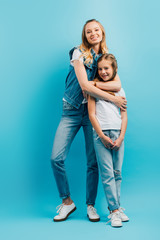 young woman in denim clothes hugging daughter wearing white t-shirt and jeans while looking at camera on blue