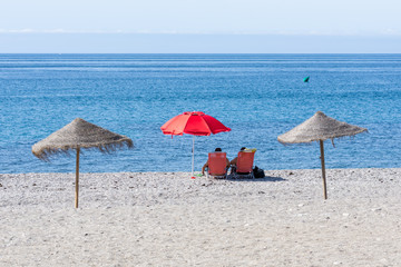 Andalusian beach a sunny summer morning with two people sitting under a red umbrella looking out to the sea