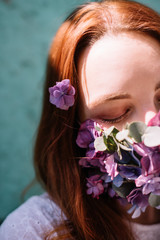 Obraz na płótnie Canvas Beautiful young redhead woman wearing a medical mask decorated with fresh blossoming purple flowers, standing on the turquoise wall background