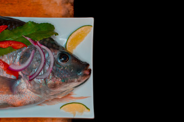 Fresh tilapia fish presented on a long plate garnished with colorful vegetables. dark food