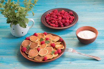 Pancake cereals and red ripe fresh raspberries in a brown plate. A bunch of Melissa in a mug on a blue wooden table. Delicious Breakfast