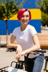 Fototapeta na wymiar Happy young woman with pink hair walks around the city on a bike in the summer. Eco-friendly mode of transport