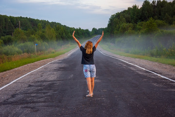Fototapeta premium A young red-haired woman looks at the road receding into the distance on a summer evening. Rural landscape with a road, trees, grass. There is fog in the background