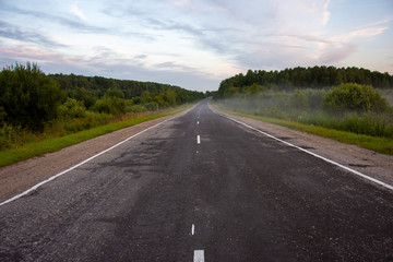 A view of the road going into the distance on a summer evening. Rural landscape with a road, trees, grass. There is fog in the background