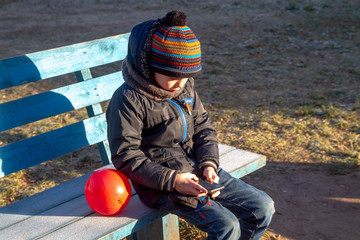 A lonely boy with a smartphone sits on a bench in winter. Frost. Cold. The boy does not play with friends, refusing to communicate with people and games.