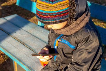 A lonely boy with a smartphone sits on a bench in winter. Frost. Cold. The boy does not play with...