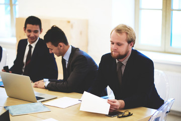 Young man successful managing director is reading paper documents with monthly report of his new project income.Businessman is sitting at the table with open portable laptop computer near his partners