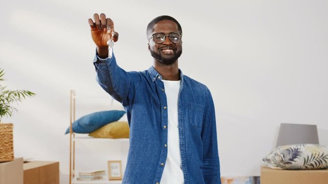 Close up of handsome african american man in denim shirt smiling while standing in new apartment. Man raises his hand up, showing bunch of keys to new house.