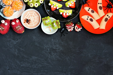 Healthy Halloween fruit snacks. Assortment of fun, spooky treats. Overhead view top border over a black stone background with copy space.