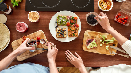 Top view of couple of vegetarians preparing various healthy toasts in the kitchen together. Man getting out sun dried tomatoes while woman adding chia seeds