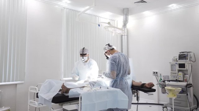 Two male surgeons at the work with time lapse effect. Action. Sedated patient lying on the table during surgical procedure inside the hospital room.