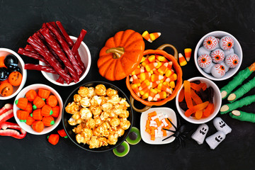 Halloween candy buffet table scene over a black stone background. Assortment of sweet, spooky treats. Top down view.