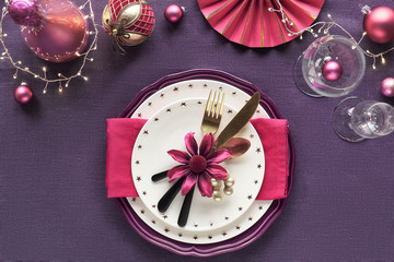 Christmas table setup with golden, pink and red decor on purple linen textile. Plates with flowers, utensils and vine glass.