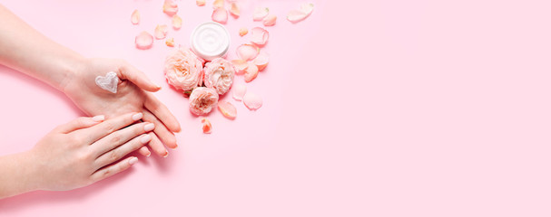 Skin care concept. Female hands with cream and roses flowers on pink background. Copy space