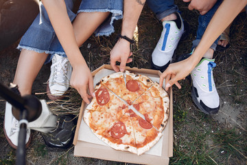 Group of friends is sitting near the lake. They wear casual clothes. Young people eat pizza outside.