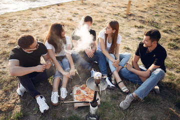 Group of friends is sitting near the lake. They wear casual clothes. Young people eat pizza outside.
