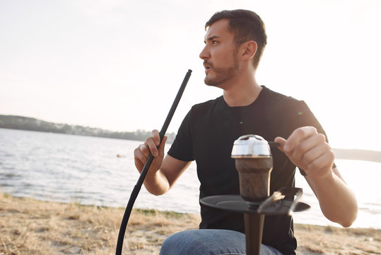 A man is fixing a hookah. He wears a black T-shirt. The man has a beard.