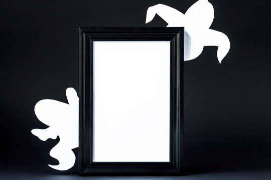 Background for Halloween. Black frame with free space on a black background. White ghostly figures peek out from behind an empty black photo frame.  
