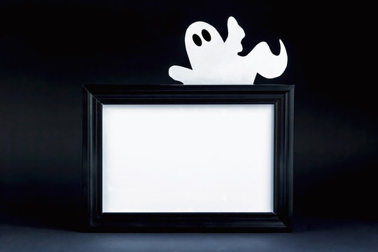 Background for Halloween. Black frame with free space on a black background. A white ghost figure peeks out from behind an empty black photo frame. Halloween typography concept. Halloween Ideas.