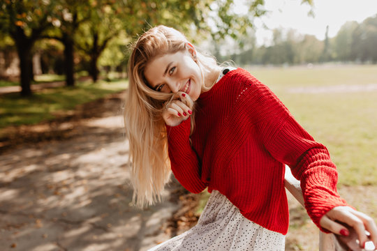 Happy young blonde in trendy red sweater and white skirt smiling in autumn park. Stylish girl with natural makeup posing outdoor.
