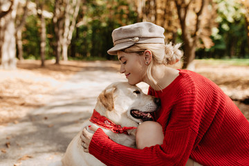 Young blonde in trendy red clothes smiling at her lovely labrador in autumn park. Lovely girl and her dog relax outdoor.