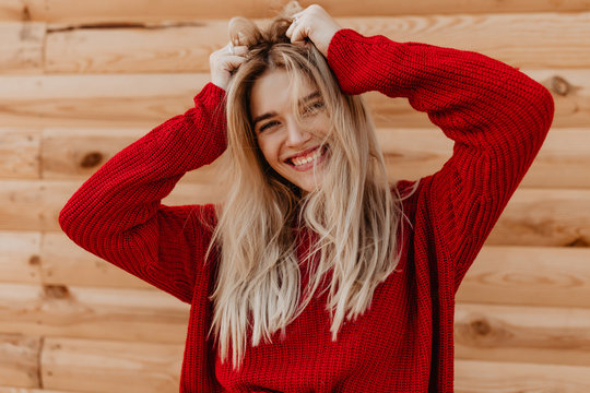Closeup photo of a stunning blonde smiling joyfully on the wooden background. Pretty girl in red sweater feeling happy outdoor.
