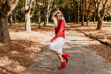 Smiling young blonde woman having fun in autumn park. Dancing in stylish red shoes and white dress.