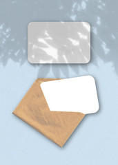 An envelope with two sheets of textured white paper on the blue background of the table. Mockup overlay with the plant shadows. Natural light casts shadows from an exotic plant