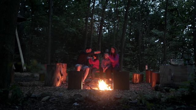 Static FHD shot of a family with two little daughters sitting on a bench by the fire in a camp at night, talking and drinking water and wine. Family enjoying their time together by the campfire.