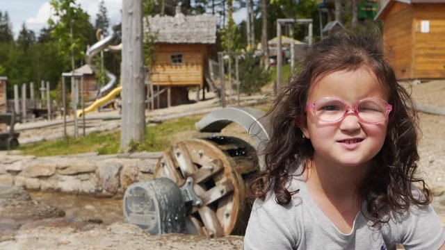 Static FHD shot of a little girl with glasses grinning and smiling at a kids playground sitting next to a small watermill and a stream with stony edges. Small girl sitting at a playground and smiling.