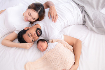 Obraz na płótnie Canvas Asian family lifestyle or mix race couple lying down on bed together relax comfortable, Asian Father with beard or Muslim dad and mother sleep hold/hug adorable newborn with love, care and protection
