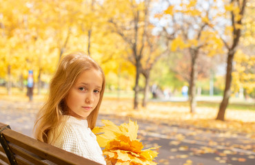 Naklejka na ściany i meble beautiful blonde teen girl sitting on a bench in an autumn Park with maple trees