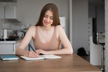 Smiling woman in listens to course using laptop at home, takes notes, young woman learns foreign languages, digital self-education,