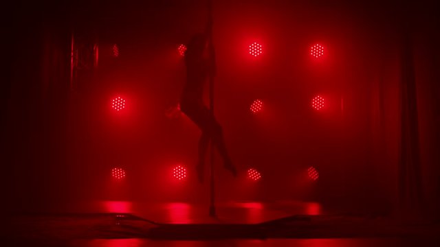 Pole dance practice. Dancer in dark studio spin on pole indoors. A silhouette looms against a smoky bright red background. Slow motion.