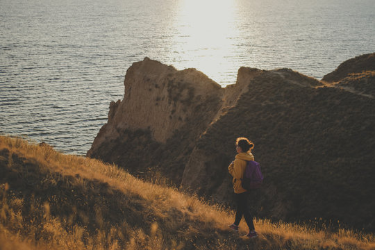 Young woman traveler with backpack in yellow hoodie walking the trail path at mountain peak, hiking to cliff by the sea during sunset, enjoying nature landscape. Soft filter. Adventure concept
