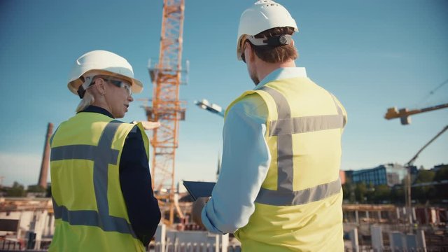 Male Civil Engineer and Young Female Building Architect Use a Tablet Computer on a City Construction Site. They Talk About the Future of Real Estate Development and Planning. Wearing Safety Hard Hats.