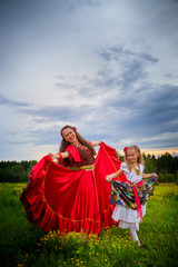 Mother and daughter in colorful dresses similar to Gypsy resting and having fun in a meadow with green grass. Models an adult woman and a little girl posing or performing in the field in the evening