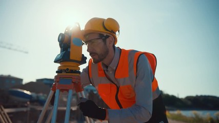 Construction Worker Using Theodolite Surveying Optical Instrument for Measuring Angles in Horizontal and Vertical Planes on Construction Site. Worker in Hard Hat Making Projections for the Building. - Powered by Adobe