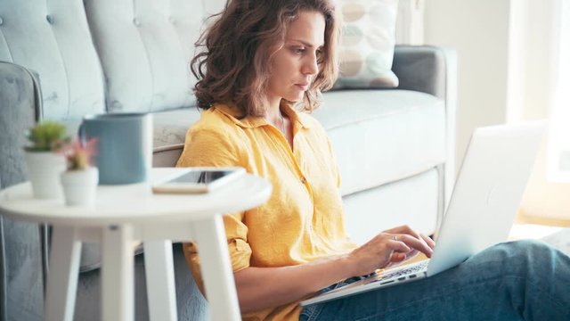Young serious woman remote employee working on her laptop at home. Freelancer at her living room working and drinking coffee from a mug.