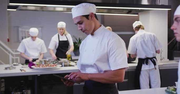 Mixed race male chef wearing chefs whites in a restaurant kitchen using a tablet and looking at came