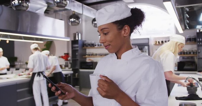 African American female chef wearing chefs whites in a restaurant kitchen using a phone and smiling