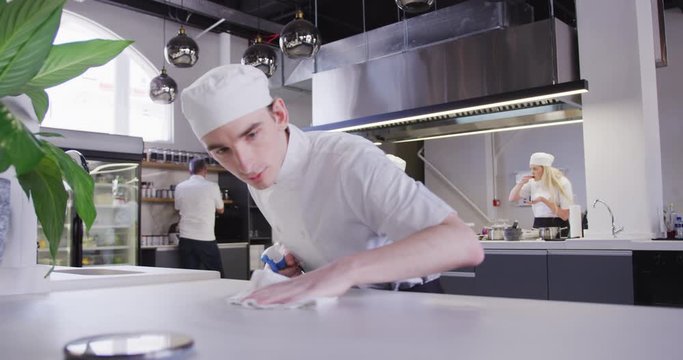 Caucasian male chef in a restaurant kitchen, cleaning a counter top, with colleagues working