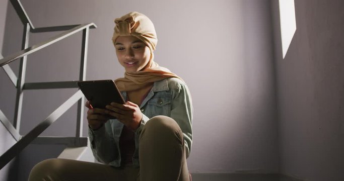 Asian female student wearing a beige hijab sitting on stairs and using a tablet