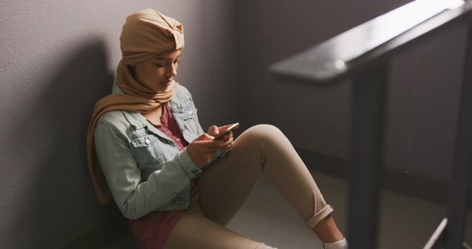Asian female student wearing a beige hijab leaning against a wall and using a smartphone