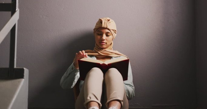 Asian female student wearing a beige hijab sitting on a staircase and reading a book