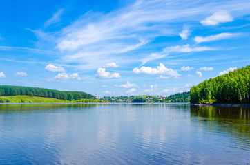 Lake in the forest on a clear summer day. A village on the shore of a reservoir. House. Settlement. Specular reflection in the water. Green foliage of trees. Coast. Blue sky with white clouds on a sun